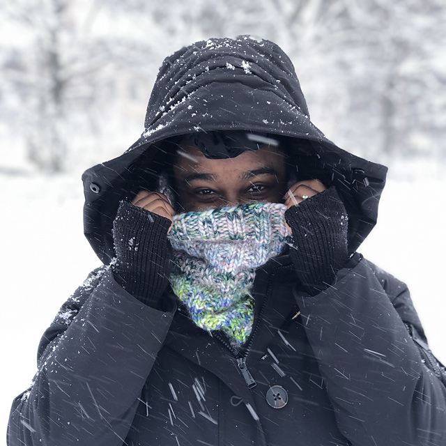 image of woman in the snow wearing a jacket and hood with a multi-color intersect cowl around her mouth and nose